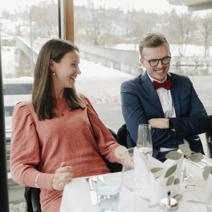 Lächelndes Paar in eleganter Kleidung, beim Essen an einem Tisch mit Ausblick.